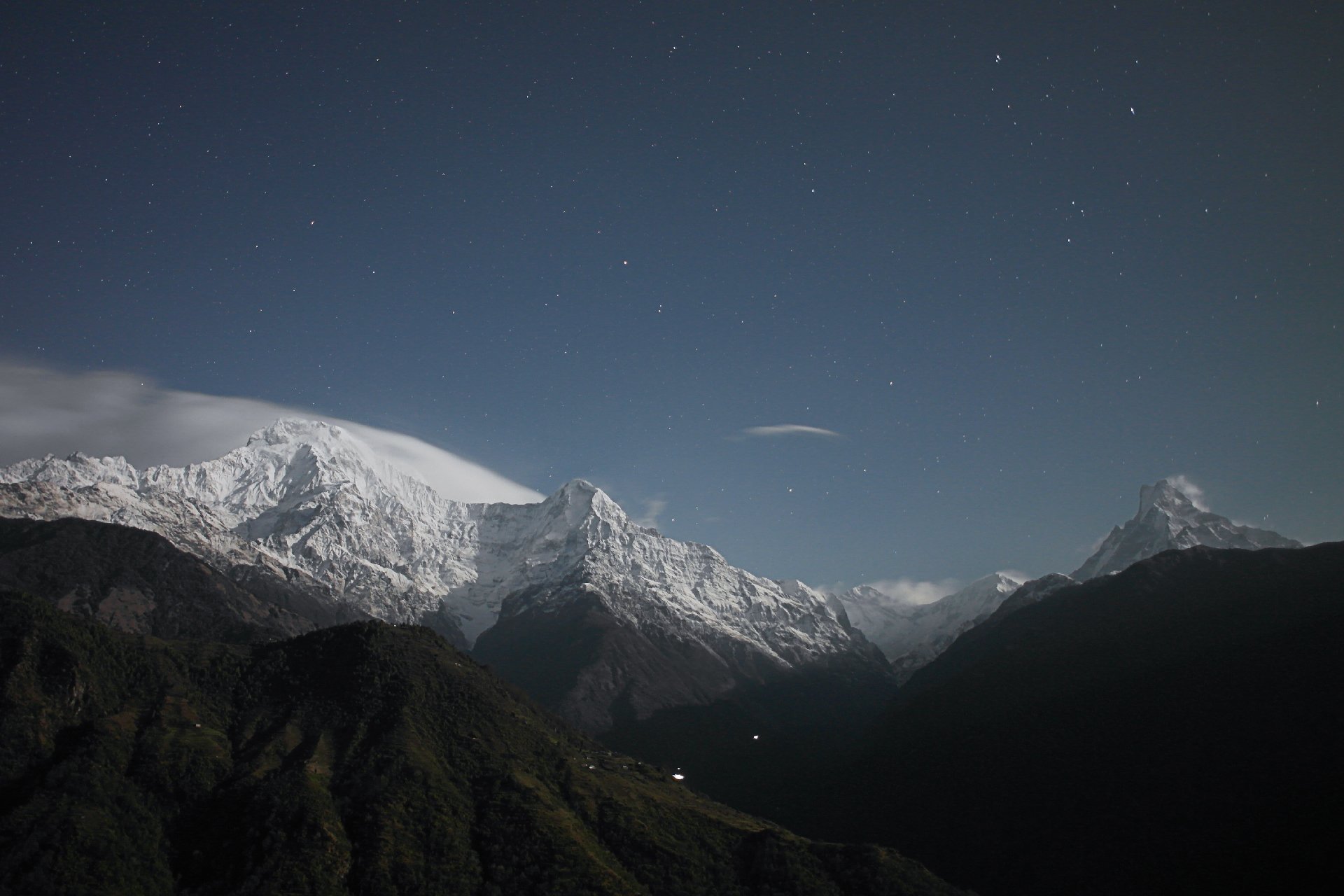 Night sky over snow-capped Himalayan peaks with a starry backdrop, blue atmosphere, and a dark green valley in the foreground under wispy clouds.