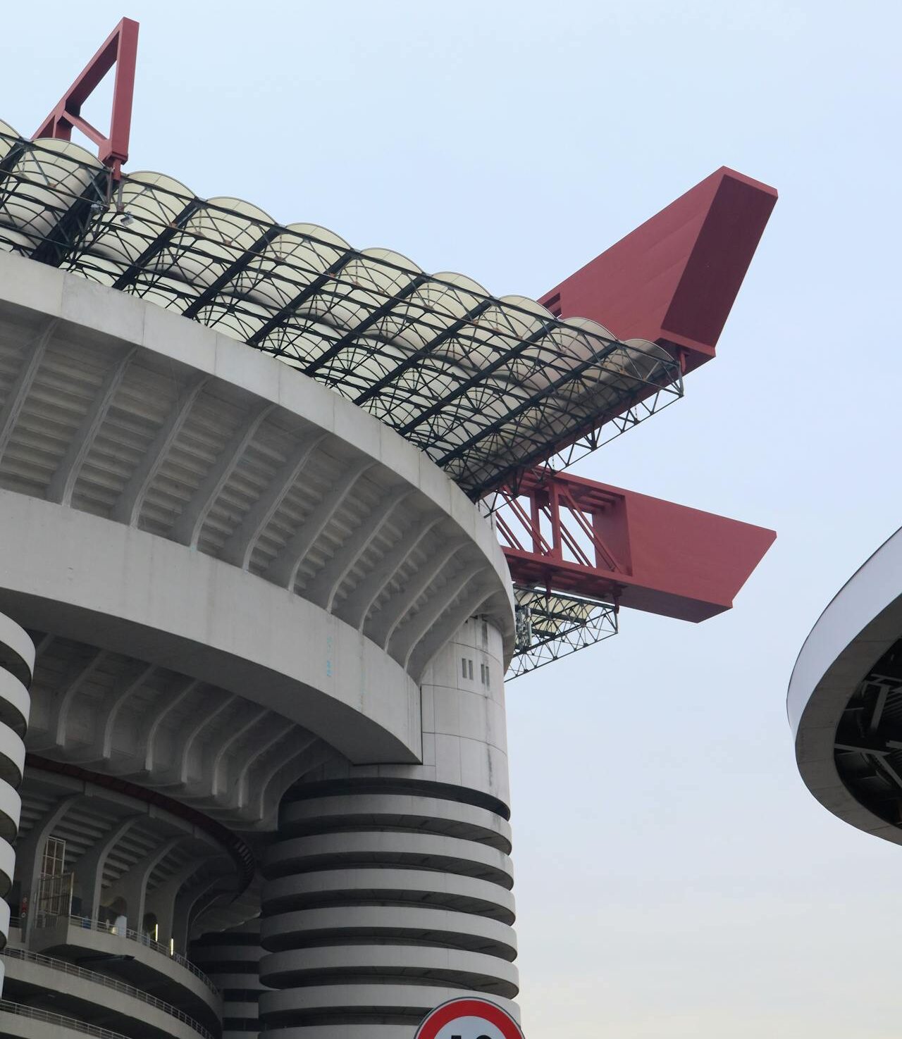Red angular structural element atop a curved concrete infrastructure with gray concrete and steel grid, photographed from below against a pale blue sky, featuring bold industrial architecture and a prominent red form