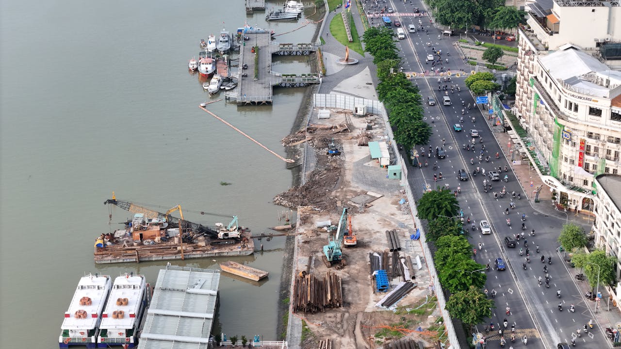 Aerial view of a riverfront construction site with cranes, barges, and stacked steel near a busy urban avenue, Vietnam skyline visible with mid-rise buildings and green trees
