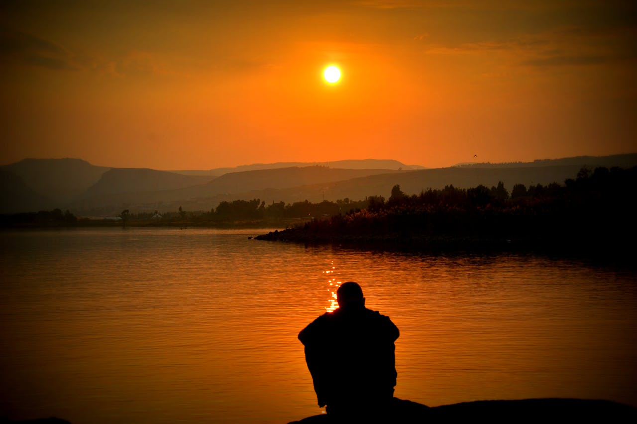 Orange sunset over calm water with a lone silhouetted figure sitting by the shore, distant hills on the horizon and a warm glow reflecting on the lake.