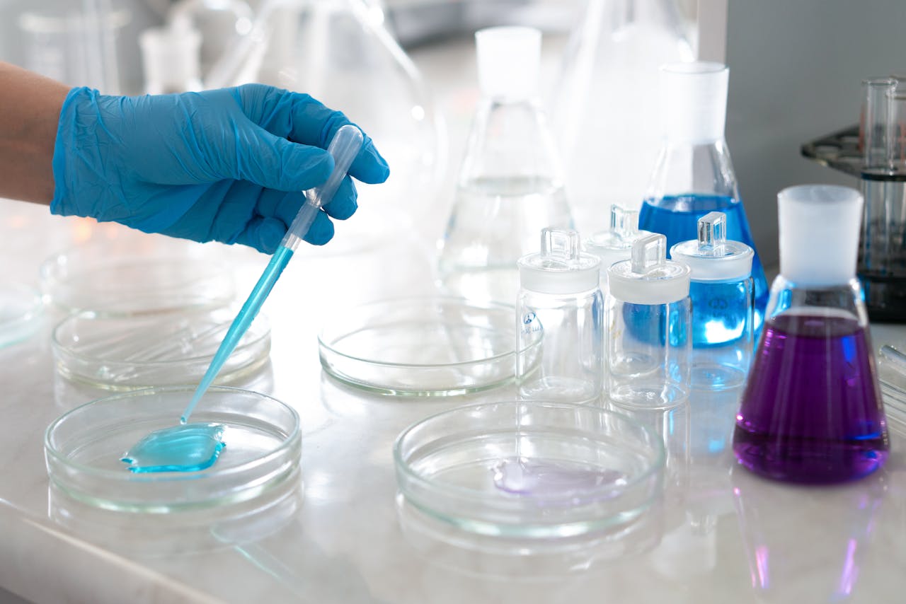 Blue-gloved hand uses a pipette to transfer turquoise liquid into a petri dish, set among lab glassware and colorful solutions in a clean laboratory workspace.
