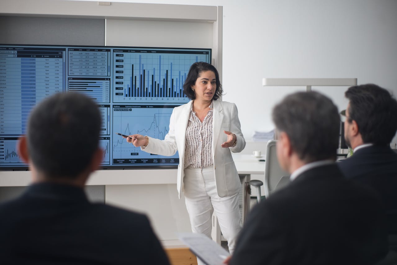 Businesswoman in a white suit presents analytics on a large multi-screen dashboard with graphs and charts to a seated group in a modern office setting.