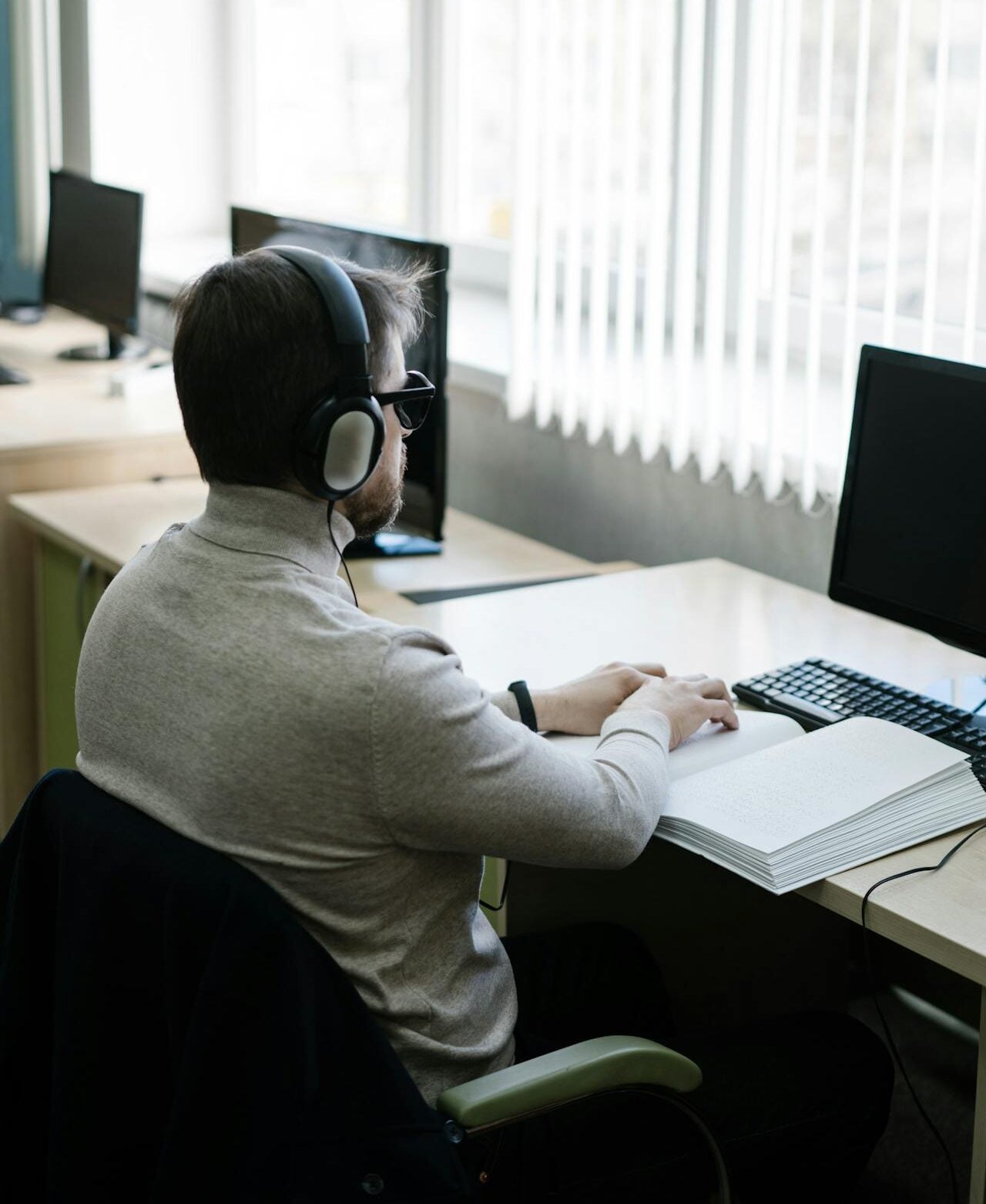Mikhail Nilov photograph of a man wearing large black headphones seated at a desk with a keyboard and notebook, in a bright office with windows and other computers in the background.