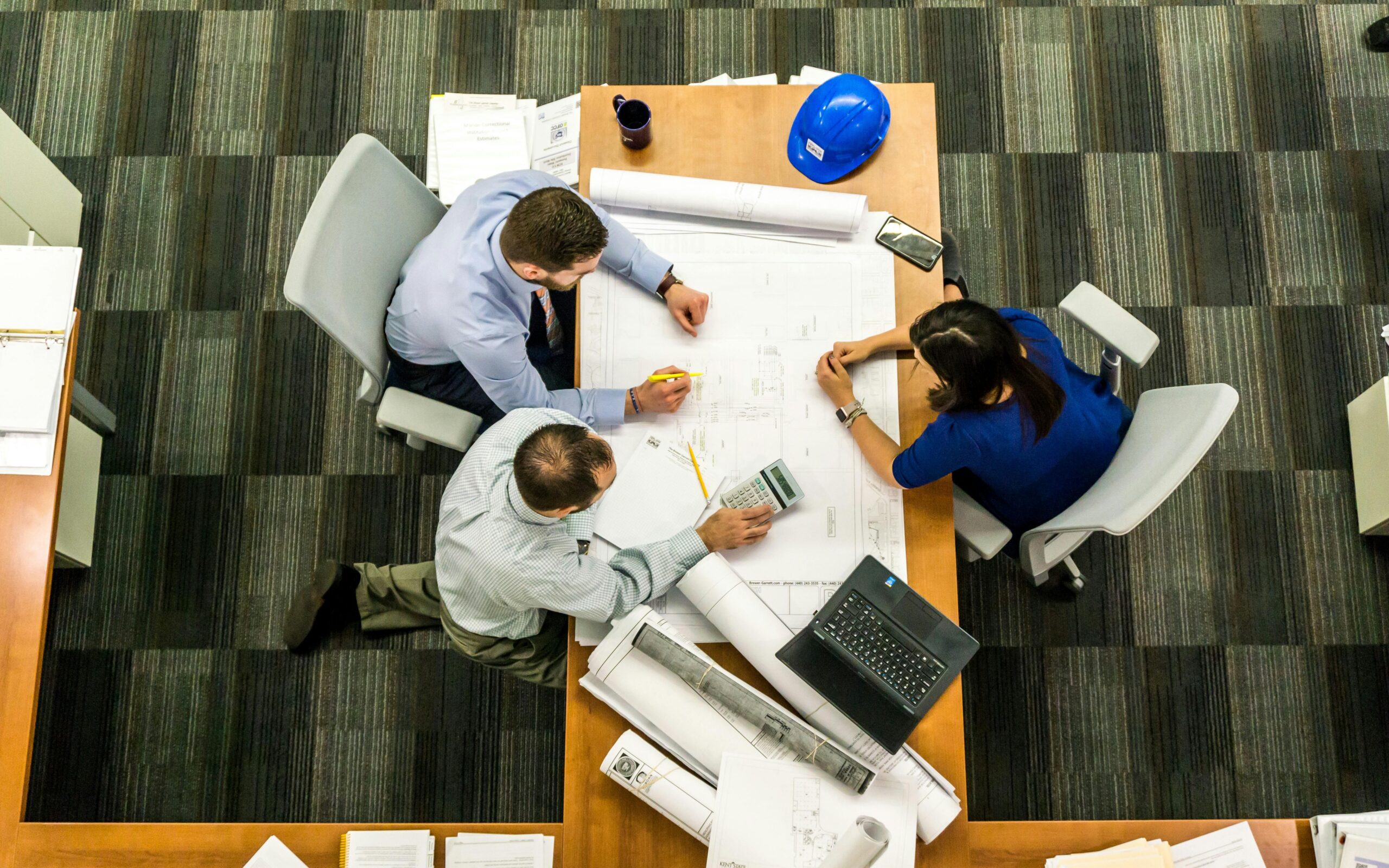 Overhead view of a blue-shirted team collaborating at a wooden desk with architectural blueprints, a blue hard hat, a smartphone, a calculator, and a laptop, highlighting dynamic planning and teamwork.
