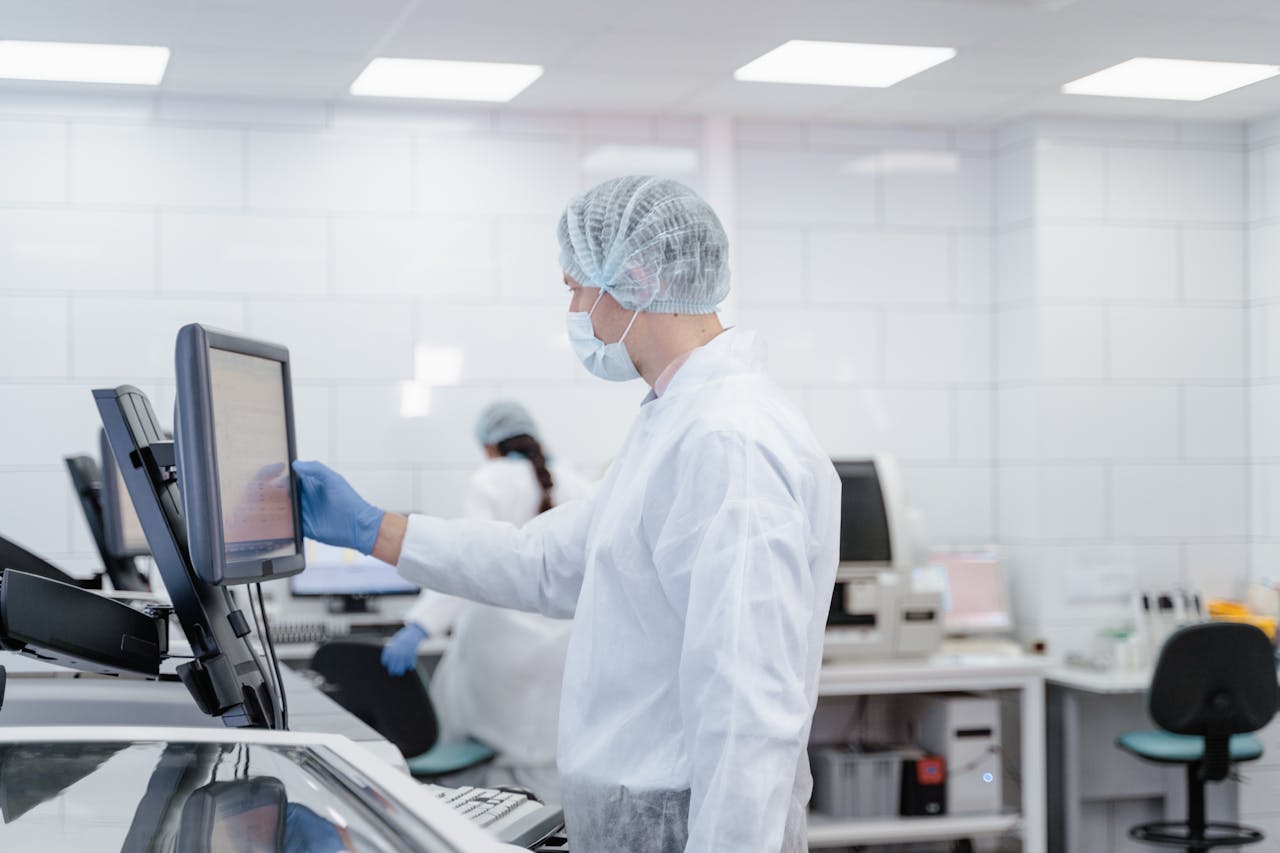 Tima Miroshnichenko wearing a white lab coat, hairnet and face mask interacts with a touchscreen monitor in a sterile medical laboratory with other technicians in the background