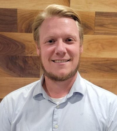 Graeme Crouch smiling in a light blue shirt against a wooden background.